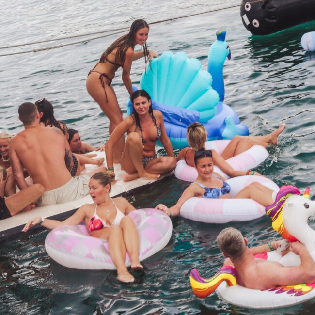 A group of people in swimsuits relax on colorful inflatable floats, including a unicorn and peacock, in the water near a boat. Some are sitting on the boat's edge, enjoying a sunny day.