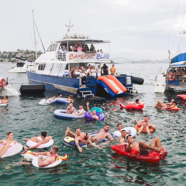 People float on inflatable tubes and loungers in the water near anchored boats, including one with a "Barefoot Blue" sign. The scene is festive, with many people smiling and enjoying a party atmosphere.