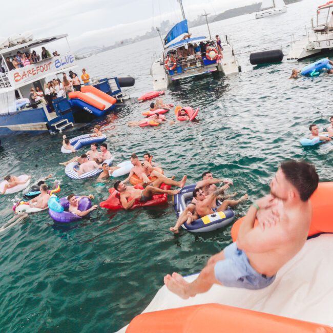 People enjoy a floating party on the water, using inflatables and slides from boats. Several boats are anchored nearby, and groups of people are swimming and lounging in the water, having fun under a cloudy sky.