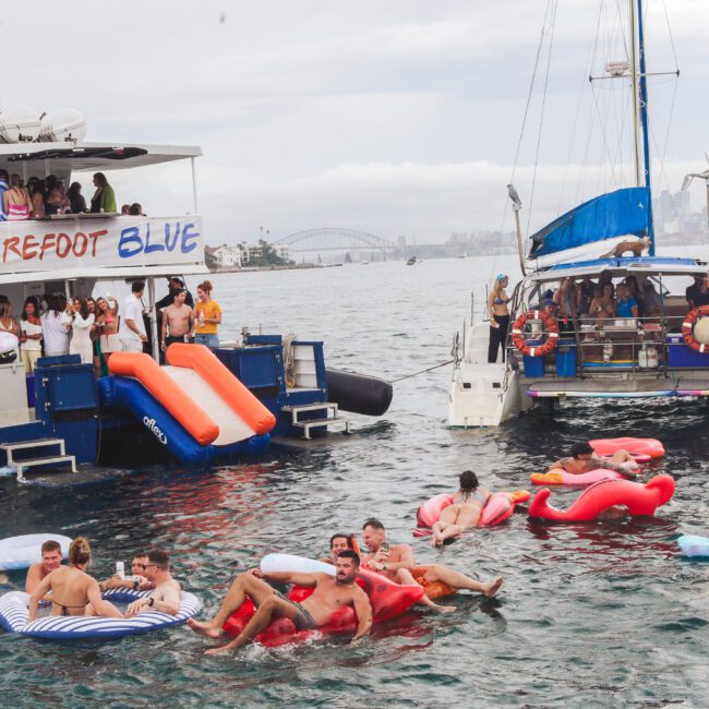 Two boats anchored close together, one labeled "Barefoot Blue," with people on board and others relaxing on colorful inflatables in the water. The scene is lively, with a city skyline and bridge visible in the background.