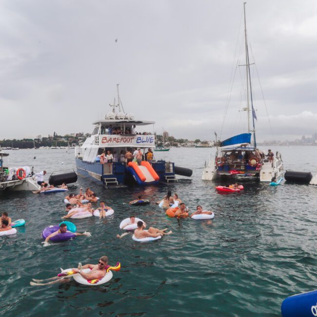 People relax on colorful pool floats in the water near several anchored boats on a cloudy day. A banner reading "BAREFOOT BLUE" hangs on one boat with inflatable slides, and the city skyline is visible in the background.