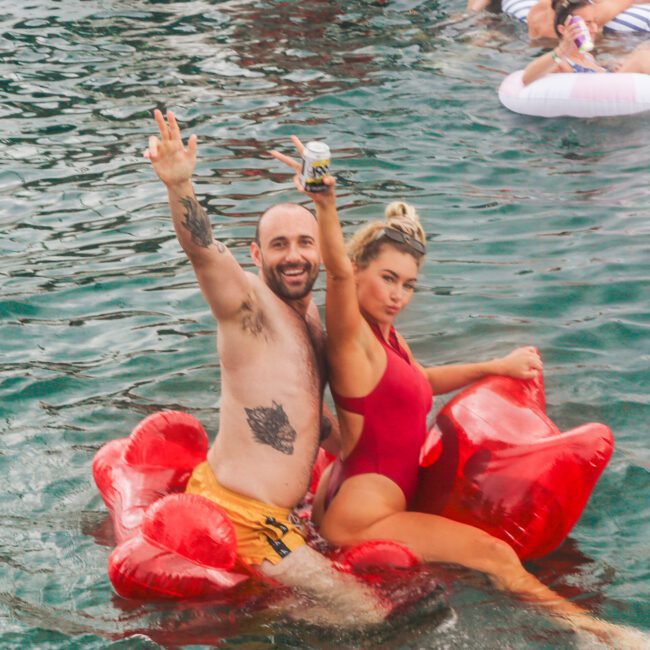 A man and a woman pose and smile on a red inflatable float in the water. The man is shirtless with yellow swim trunks, and the woman wears a red swimsuit. Other people are relaxing on inflatables nearby.