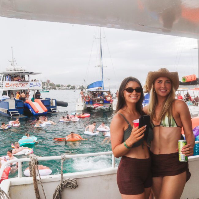 Two smiling women in swimsuits stand on a boat holding drinks, with one wearing sunglasses and the other a straw hat. In the background, people swim and relax on floaties near another boat labeled "Barefoot Blue.