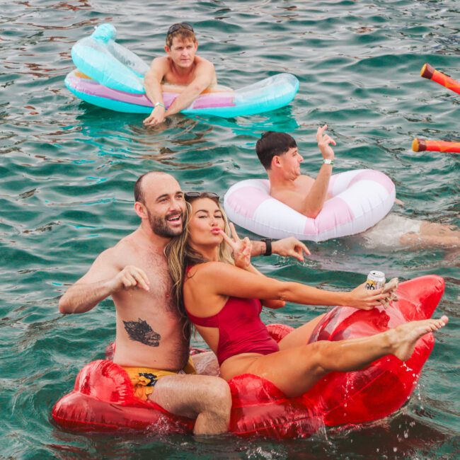 Four people float in a body of water on inflatable pool toys. A smiling man and woman pose together in front; the woman holds a drink. Two other men float behind them on separate inflatables, one shaped like a unicorn.