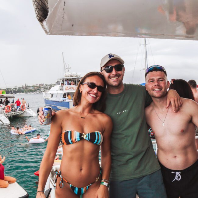 Three friends, two men and one woman in swimwear and sunglasses, smile and pose together on a boat during a lively party on the water with other boats and people in the background.