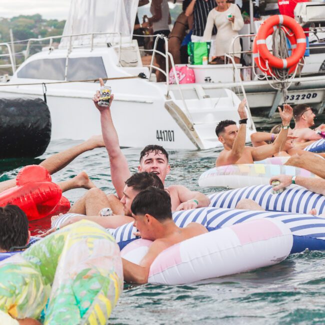 A group of people relaxing on colorful inflatable pool floats in the water near a docked catamaran boat, some holding drinks and smiling, with a lively atmosphere.