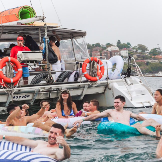 People relax and socialize on inflatables in the water near a boat, where others stand and watch. The boat is anchored close to shore with buildings and greenery in the background.