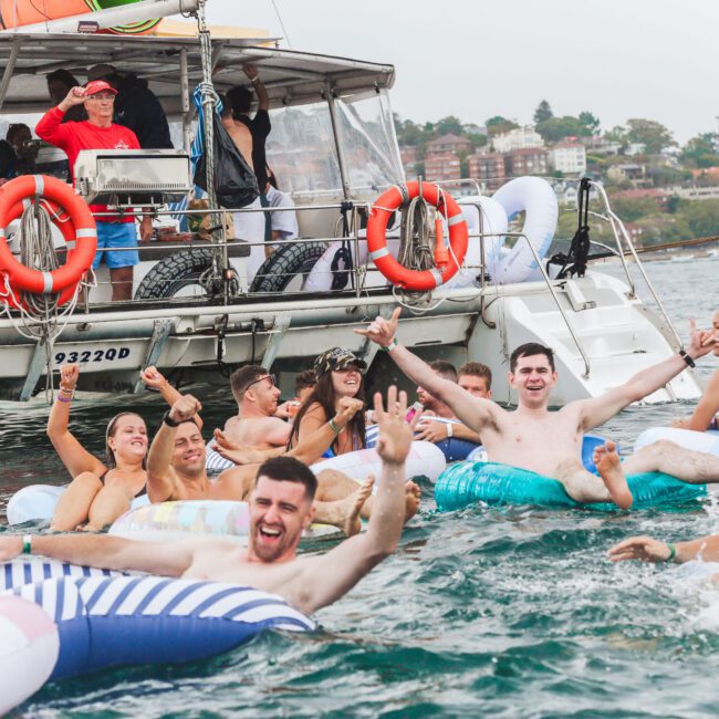 A group of young adults in swimwear laugh and wave while floating on inflatable pool toys in the water near a boat filled with more people, with buildings and greenery visible in the background.
