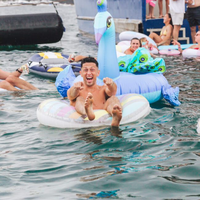 A group of people laugh and relax on inflatable pool floats, including a large blue peacock float, in the water near a dock on a sunny day.