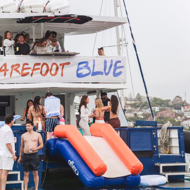 A group of people socialize on the deck and lower level of a boat named "Barefoot Blue," which features an inflatable slide and floaties at the back, cruising near a coastline with houses visible in the background.