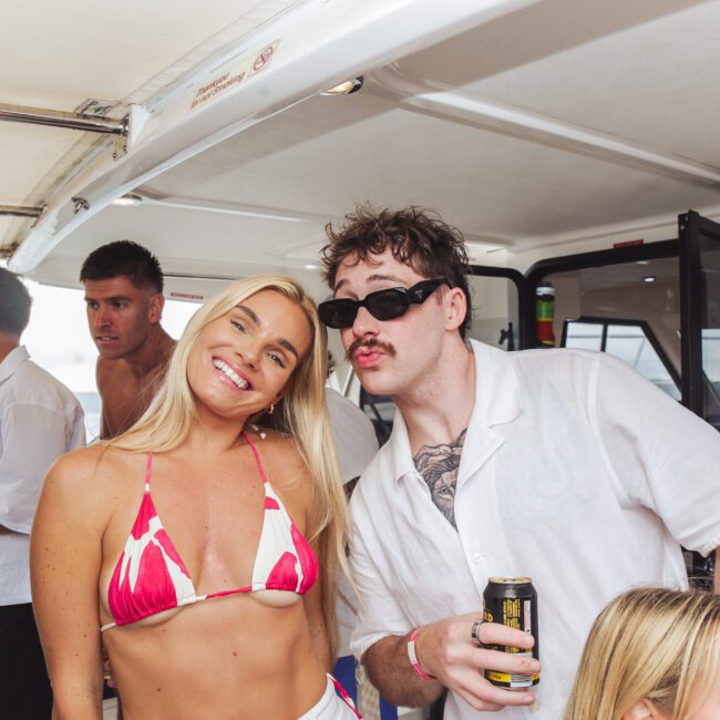 A group of young adults relax on a boat. A smiling woman in a pink and white bikini poses next to a man in sunglasses and a white shirt holding a drink. Others socialize in the background.