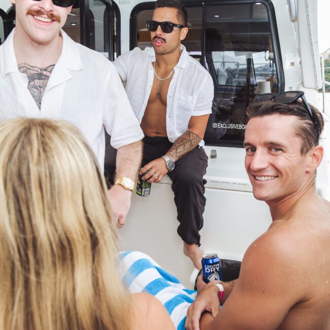 Four people are relaxing on a boat. Two men in open shirts are sitting, one holding a can. A shirtless man smiles at the camera, and a woman with long blonde hair sits with her back to the camera, wrapped in a towel.