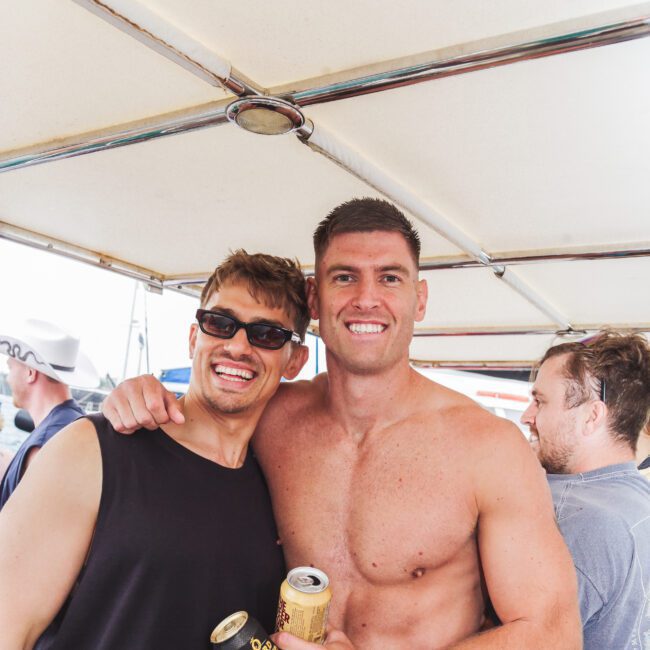 Two smiling men pose together on a boat, one wearing sunglasses and a black tank top, the other shirtless and holding canned drinks. Other people are visible in the background under a canopy.