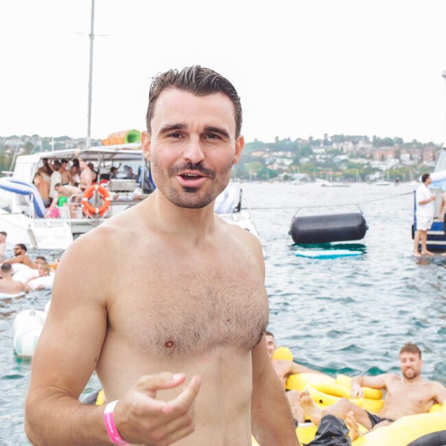 A shirtless man with dark hair stands in the foreground at a lively floating boat party, with people relaxing on inflatables and boats in the background on a sunny day.