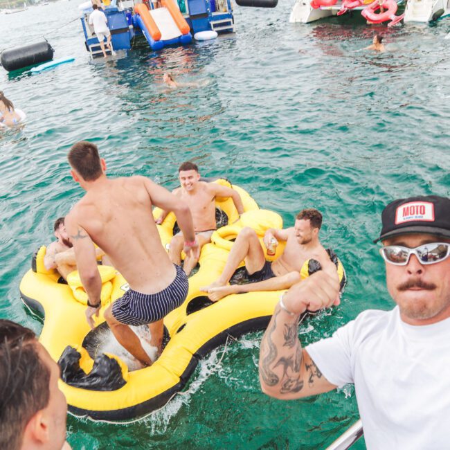 A group of young men in swimsuits enjoy a party on a yellow inflatable float in the water, surrounded by boats and people. A man in sunglasses and a white cap poses and flexes his arm at the front.