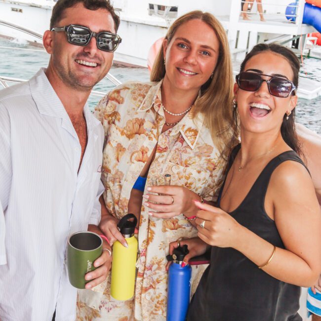 Three people smiling and posing on a boat. The man on the left wears sunglasses and a white shirt, the woman in the middle wears a floral outfit, and the woman on the right wears dark sunglasses and a black dress.