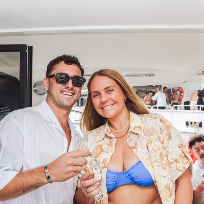 A man and woman smile at the camera while holding drinks on a boat. The woman wears a blue bikini and a patterned shirt, the man wears sunglasses and a white shirt. People are visible in the background enjoying the boat party.