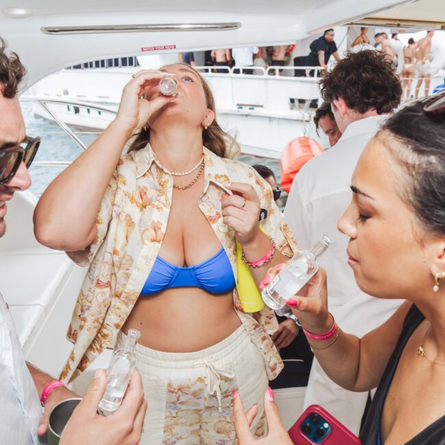 Three people on a boat are enjoying drinks; one woman in a blue bikini top tilts her head back to take a shot, while another woman and a man hold bottles, surrounded by a lively group and water in the background.