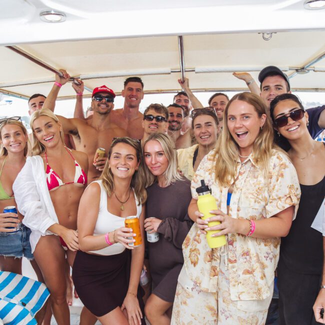 A group of people, smiling and posing together on a boat. Most are wearing casual summer clothes or swimwear, holding drinks, and appear to be enjoying a lively gathering or party.