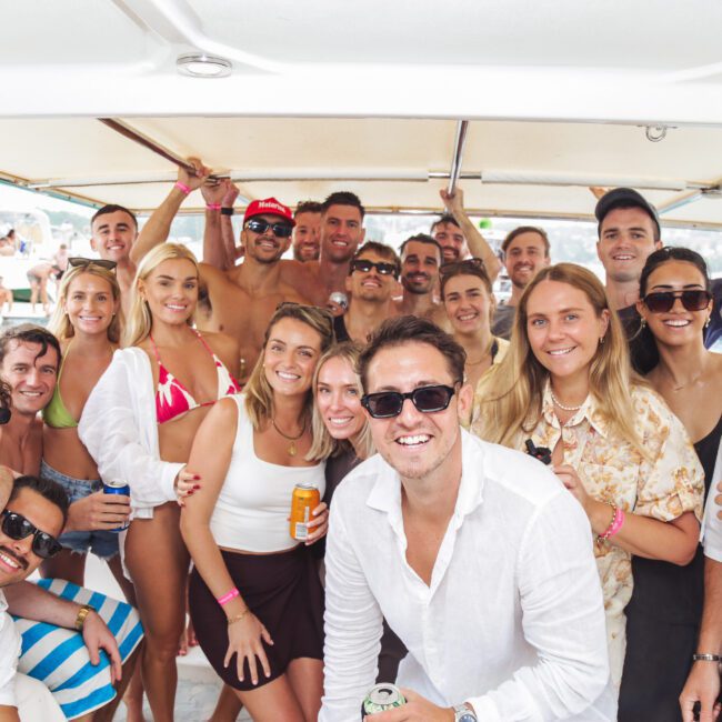 A group of smiling young adults pose together on a boat, enjoying a sunny day on the water. Many wear sunglasses and light summer clothing, holding drinks and looking relaxed and happy. Other boats are visible in the background.