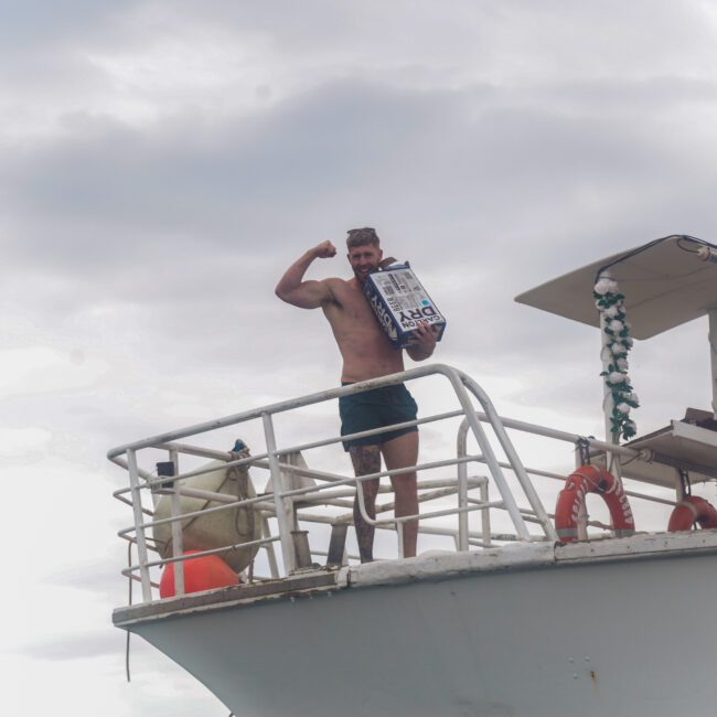 A man in swim trunks stands on the deck of a boat flexing his arm and holding a box. The sky is cloudy, and another person in sunglasses is partially visible, making a peace sign.