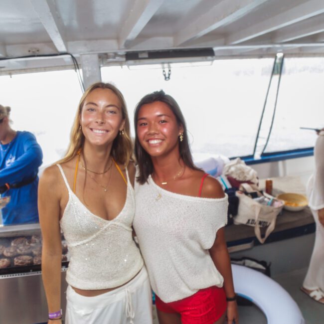 Two women smile and pose for a photo on a boat, with other people in the background. One woman wears a white sparkly top, and the other wears a white top with red shorts. There is a grill with food and bright daylight outside.