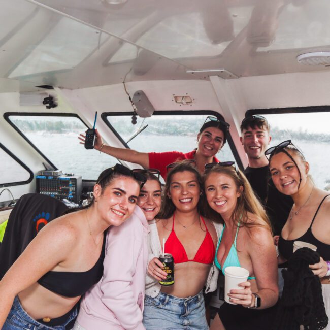 A group of seven young adults in swimwear smile and pose together on a boat. Some hold drinks, and a lake or river is visible through the windows behind them. The mood is cheerful and relaxed.