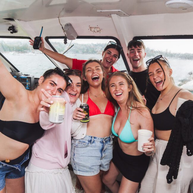 A group of happy young adults in swimsuits and summer clothes smile and pose with drinks on a boat, celebrating together with water visible through the windows behind them.