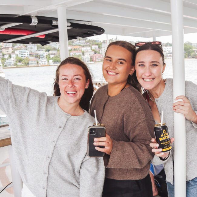 Three women smiling and holding drinks stand on a boat. They are dressed casually in shorts and sweaters, with water and houses visible in the background. The scene looks cheerful and relaxed.