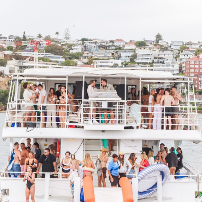 A large group of people party on a multi-level boat near the shore, with many wearing swimsuits. The background shows waterfront houses on a hillside. The scene is lively and festive.