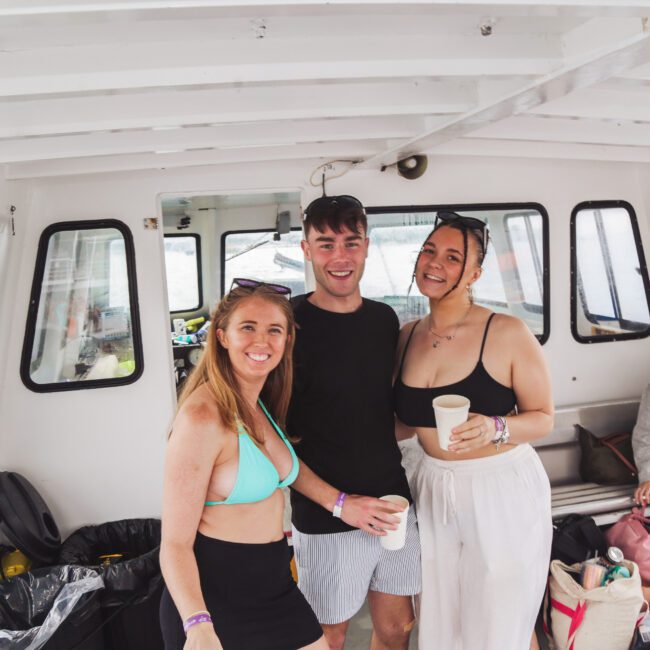 Three people smile at the camera on a boat, holding drinks. The group includes two women in summer outfits and a man in a black shirt and shorts. The boat is on the water, with bags and belongings nearby.