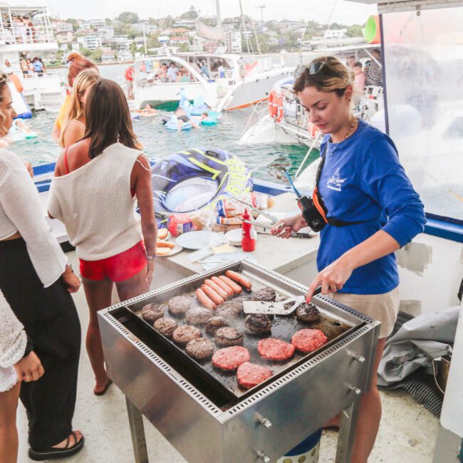 A woman grills burgers and hot dogs on a boat while several people wait in line. Other boats and people in swimsuits are visible in the background on the water.