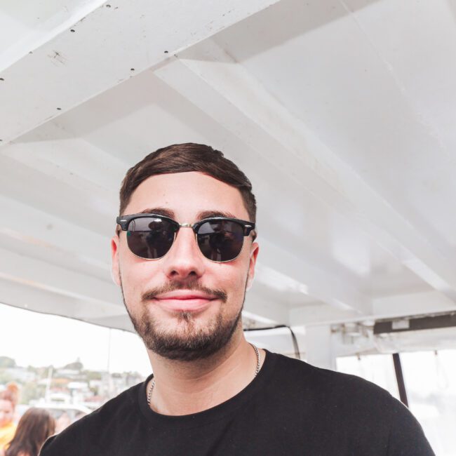 A man wearing sunglasses and a black shirt smiles at the camera while standing under the roof of a boat, with blurred people and scenery visible in the bright background.