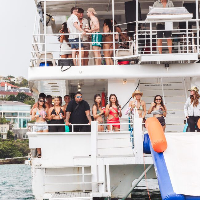 A group of people in swimwear are socializing on the deck of a white party boat with drinks in hand. The boat has an inflatable slide and is docked near waterfront houses.