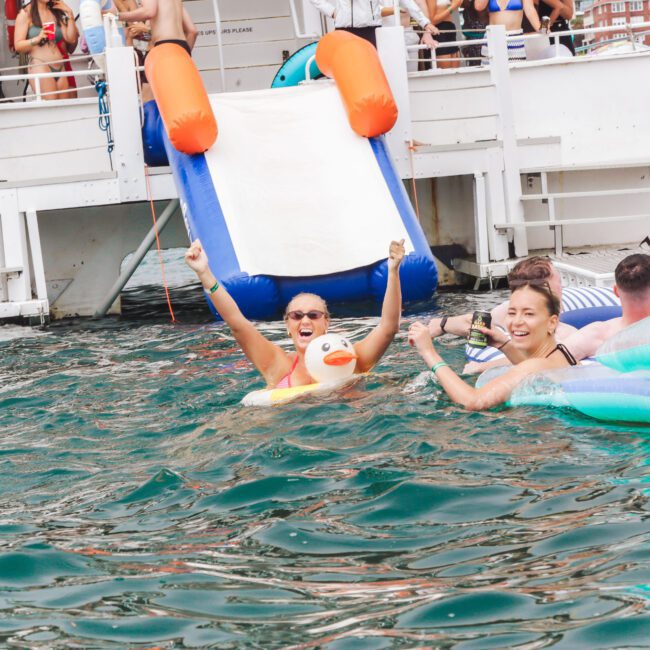 Two smiling women in swimsuits float on inflatable pool toys in the water near a boat with a slide, surrounded by people enjoying a sunny day.