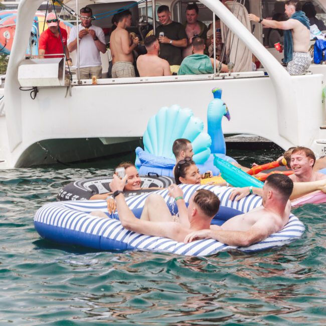 A group of people relax on inflatable floats in the water near a catamaran. Some socialize on the boat while others lounge and play in the water, enjoying a sunny day. The scene looks lively and festive.