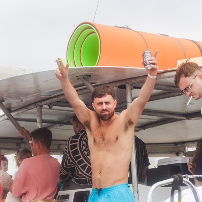 A shirtless man with arms raised holds a can and smiles on a crowded boat. Others around him chat or look away. Rolled-up mats are on the roof, and the mood appears casual and festive.