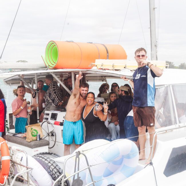 A group of people cheer and smile on a catamaran boat, some in swimsuits, with pool floats, mats, and drinks. One man holds a tray of food. The boat is near the coast on a cloudy day.