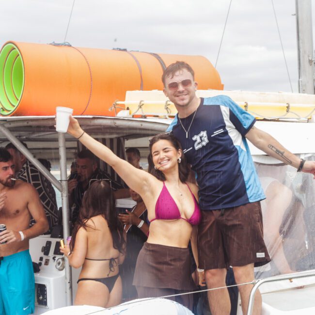 A group of young adults in swimsuits enjoy a party on a boat. A smiling woman in a pink bikini and a man in a blue shirt stand at the front, raising their drinks. Others mingle in the background under cloudy skies.