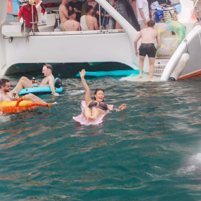 A group of people enjoy swimming and lounging on inflatables near a white boat. Some are in the water, while others are on the boat. Everyone looks relaxed and happy, celebrating in a lively, social atmosphere.