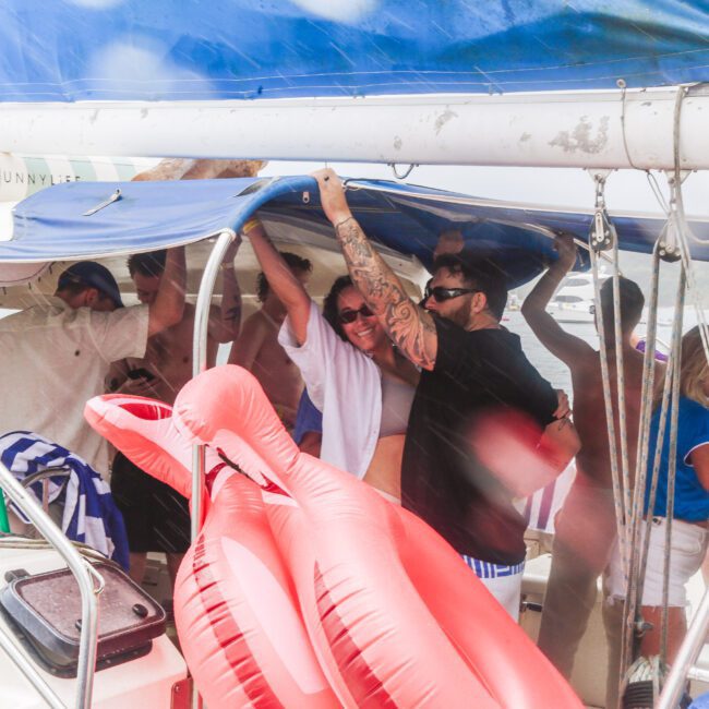 A group of people, some wearing sunglasses, gather under a boat canopy during rainy weather. A large inflatable red flamingo floats in the foreground. The mood appears lively despite the wet conditions.