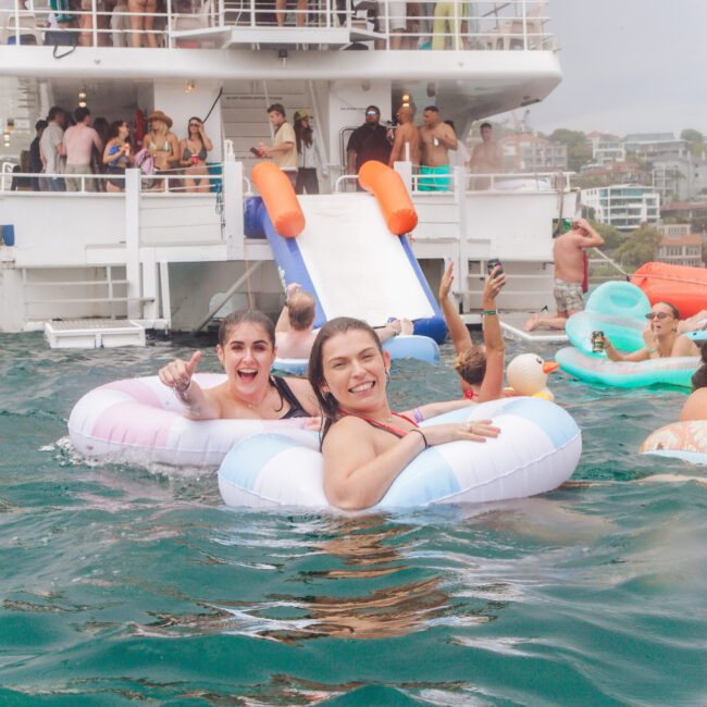 Two women in swimsuits float on inflatable rings, smiling at the camera, in the water near a large boat with a slide and many people enjoying a party. Others swim and take photos in the background.