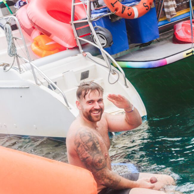 A shirtless man with tattoos sits at the edge of a boat, smiling and waving while partially submerged in water. Inflatable rings and boating equipment are visible in the background.