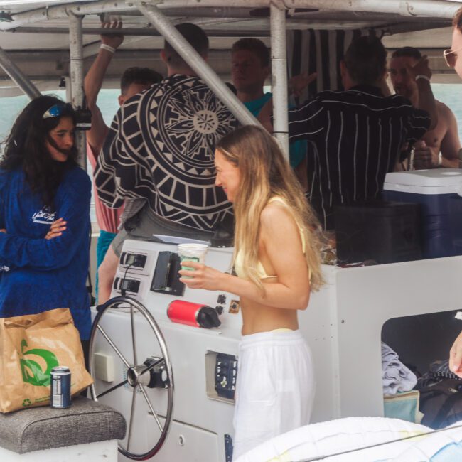 A group of people socializing on a boat, some holding drinks. A woman in a blue shirt talks to another woman in a yellow top. The scene is relaxed, with snacks and drinks on a counter next to the boat’s steering wheel.