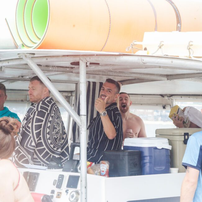 A group of people, some in swimsuits, gather and smile on a boat under a canopy. Bright light highlights the relaxed, cheerful atmosphere. Rolled-up mats and coolers are visible in the background.