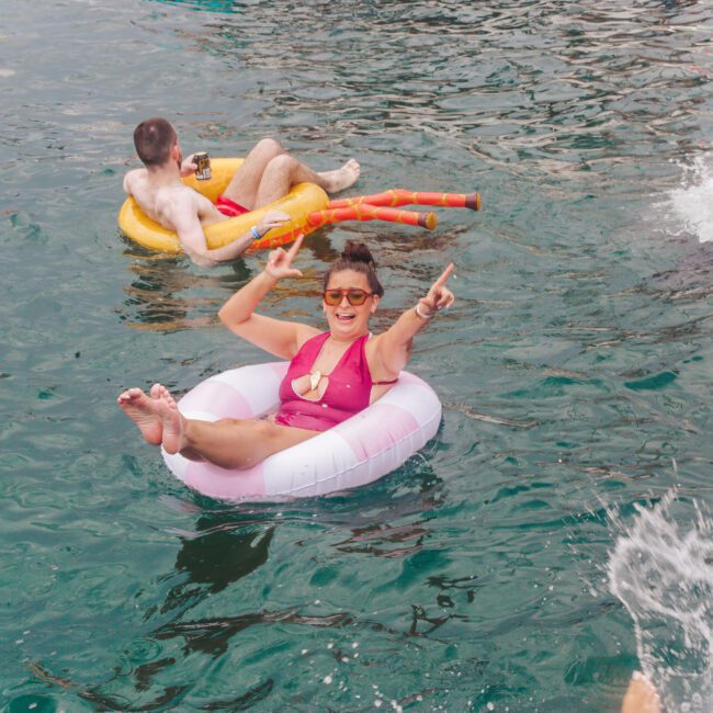A woman in a pink swimsuit and sunglasses floats on a swim ring, smiling and pointing while making a peace sign. Other people relax and splash in the surrounding water.