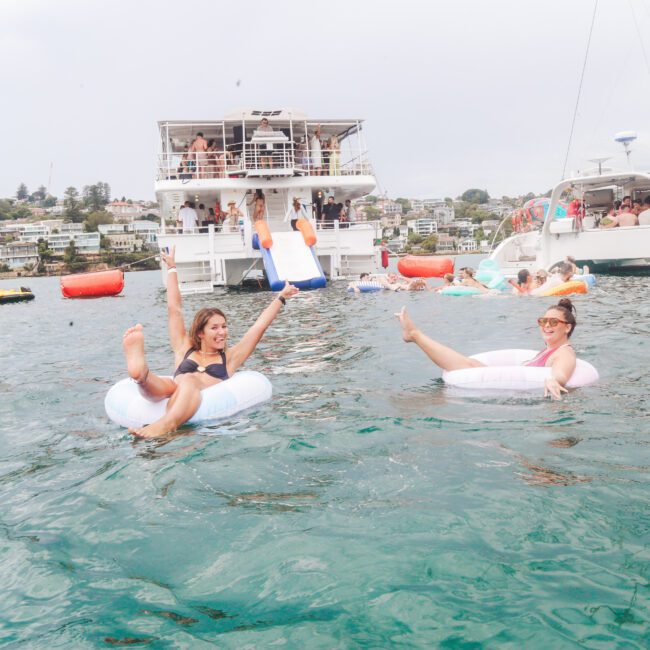 Two women smile and raise their arms while floating on inflatable rings in the water, near boats and catamarans, with more people and city buildings visible in the background.