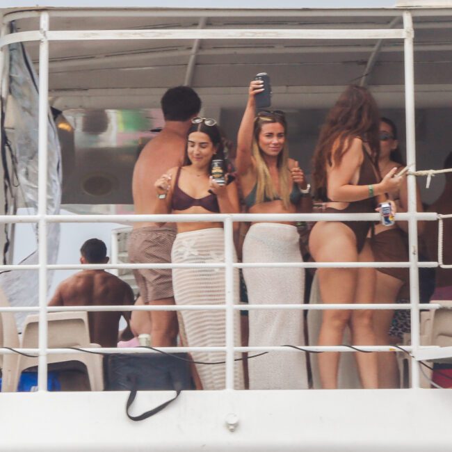 A group of people in swimsuits are standing and holding drinks on the deck of a white boat, enjoying a party. Some are facing the camera, others are talking or looking away. The background shows part of the boat’s interior.