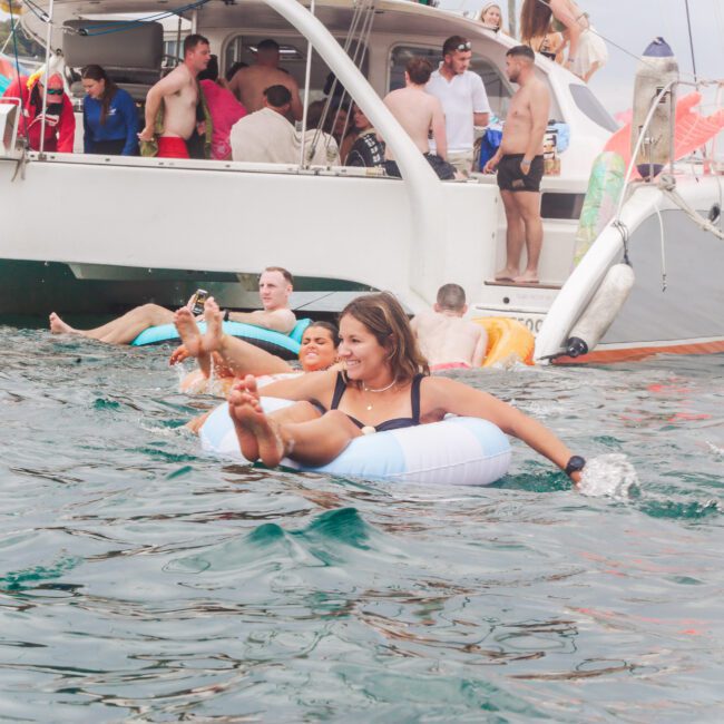 People relax and have fun on a boat and in the water; a woman on a float ring smiles with others around her, while more people stand and mingle on a large catamaran in the background.