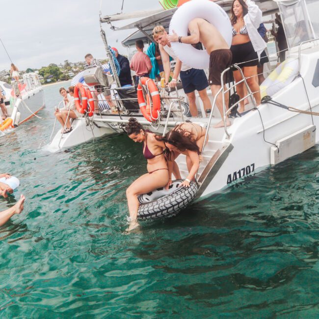 A group of people in swimsuits enjoy a boat party; two women climb onto a boat using a tire, while others relax on inflatables in the water. Several people stand and watch from the boat. The weather is cloudy.